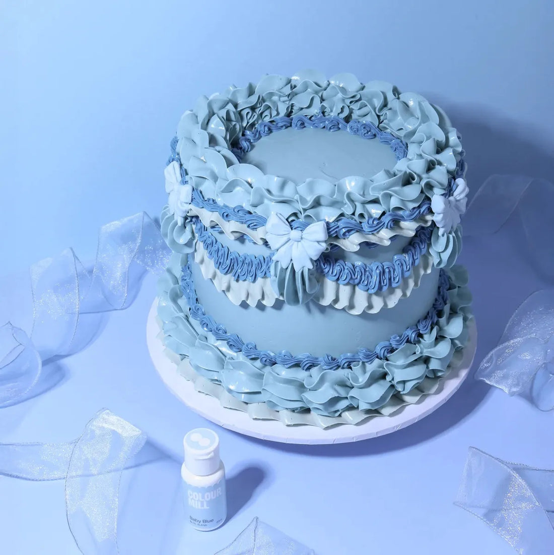 Elegant blue cake with intricate ruffled icing and bows on a light blue background. Includes a small "baby blue" food coloring bottle, creating a serene mood.