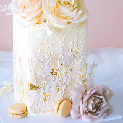 A decorative cake featuring intricate white detailing and gold accents. It has a smooth surface topped with pale rose flowers and small leaves. At the base, there are two macarons and a light pink rose. The background is softly blurred, emphasizing the cake and its embellishments.