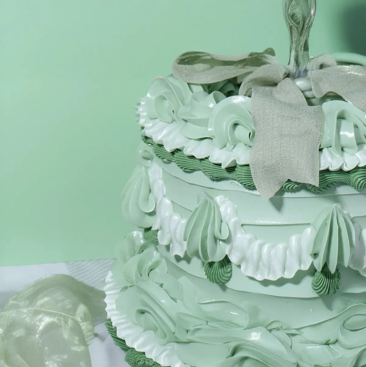 Pale green, intricately decorated cake with white frosting swirls and frills. A silver bow sits on top, creating a festive, elegant appearance.