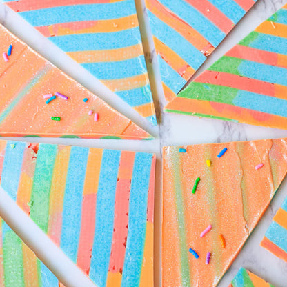 The image shows a beautifully arranged plate of colorful, triangular slices of a dessert. Each slice features a vibrant pattern of alternating stripes in shades of sky blue, pink, orange, and green. Some slices are adorned with small, colorful sprinkles scattered on top, adding to the festive and playful appearance. The background is a light-colored surface, highlighting the bright colors of the dessert slices.