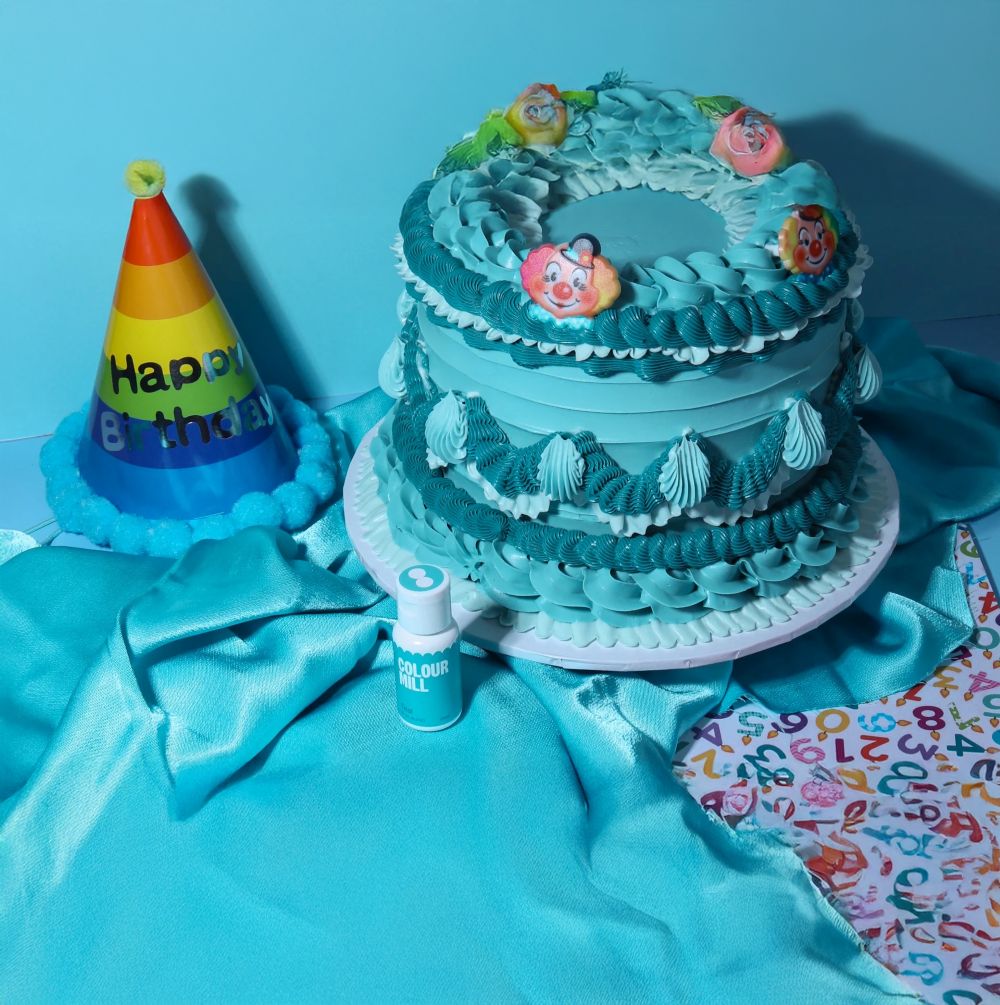 A blue two-tiered cake with colorful piped clowns, next to a rainbow birthday hat and a teal cloth. Festive, vibrant, and playful atmosphere.