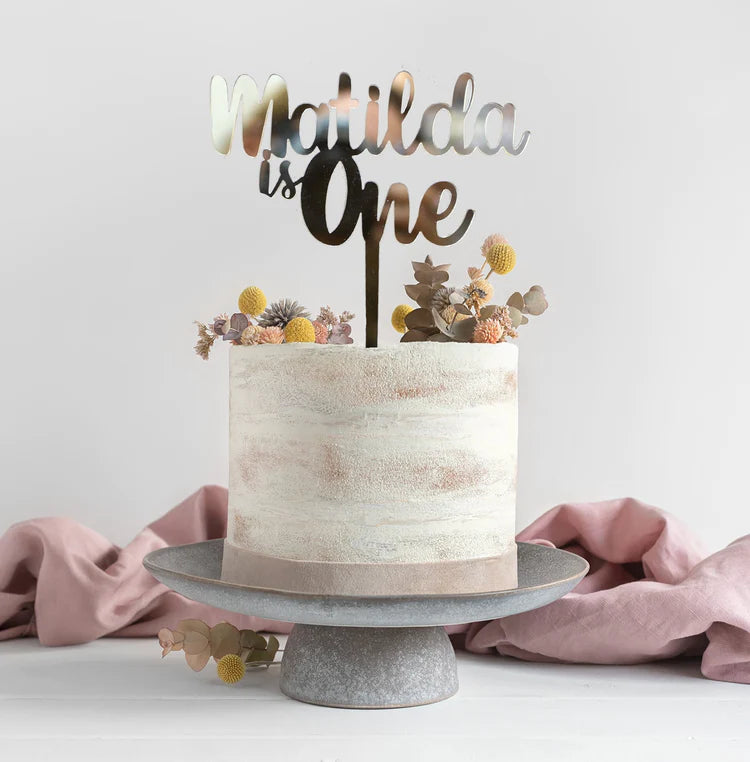 A decorated cake sits atop a round, gray cake stand. The cake has a smooth, pale frosting with subtle pink and beige streaks. Atop the cake is a shiny acrylic cake topper that reads &quot;Matilda is One&quot; in a combination of elegant fonts. The cake is adorned with small, colorful flowers in shades of yellow, purple, and green, along with some greenery, adding a festive touch. A soft, blush-colored cloth drapes gently in the background.