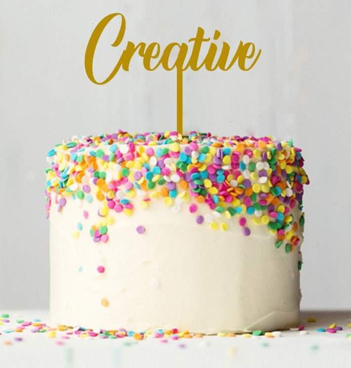 A round cake with white frosting is topped with colorful sprinkles in various shapes, including pink, yellow, blue, and purple. A gold card stock cake topper reading "Creative" is inserted into the center of the cake. The background is a soft, neutral color, emphasizing the cake&