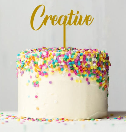 A round cake with white frosting is topped with colorful sprinkles in various shapes, including pink, yellow, blue, and purple. A gold card stock cake topper reading "Creative" is inserted into the center of the cake. The background is a soft, neutral color, emphasizing the cake&