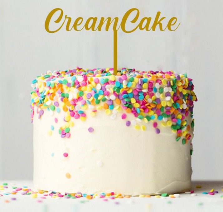 A round cake covered in white frosting with colorful sprinkles on top. A gold card stock cake topper reading "Cream Cake" is placed on the center of the cake. The scene is bright and cheerful, with additional sprinkles scattered on the surface around the cake.