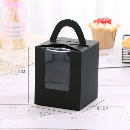 A black cardboard cupcake box with a handle, designed to hold a single portion. The box measures 11 cm in height and 9.2 cm in width. It features a clear window on the front, allowing visibility of the cupcake inside. In the background, there is a decorative cupcake topped with red fruit and a plate of cookies. The setting includes a light-colored wooden surface and some decorative crystals scattered around.