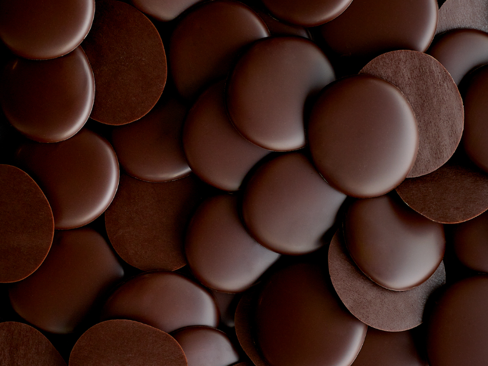 A close-up image of an assortment of dark chocolate discs, arranged in a scattered pattern. The chocolate discs vary slightly in size and texture, showcasing a rich dark brown color with a glossy finish.