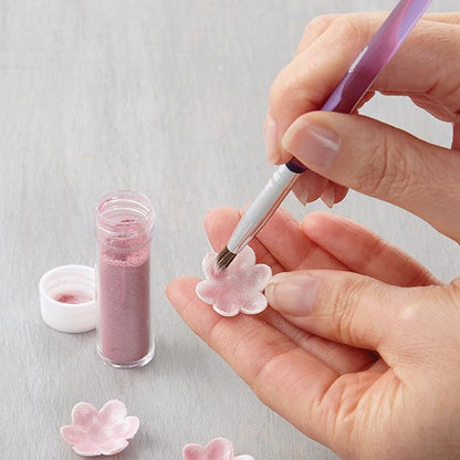 A close-up image shows a person using a decorating brush to apply pink powder to a small flower-shaped decorative piece. A container of pink powder is visible alongside another small lid. The background is a light-colored surface.
