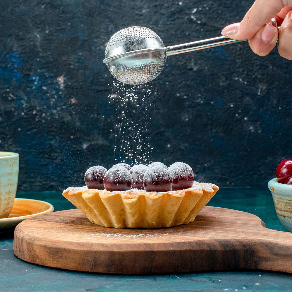 A hand dusts powdered sugar over a cherry-topped tart on a wooden board. The scene exudes a cozy, inviting vibe with a dark, textured background.