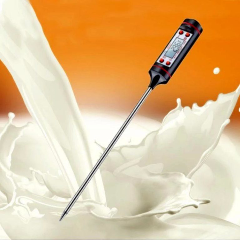 A digital candy thermometer with a long stainless steel probe is shown above a splash of white liquid resembling milk. The thermometer has a black body with red accents and a digital display indicating the temperature. The background is a gradient of orange, creating a warm ambiance.
