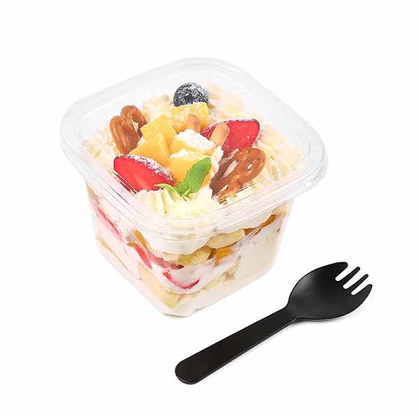 A clear square plastic dessert cup filled with layered dessert featuring whipped cream, strawberries, pineapple chunks, and decorative toppings. A small black plastic spoon is placed next to the cup. The lid of the cup is transparent, allowing visibility of the colorful layers inside.