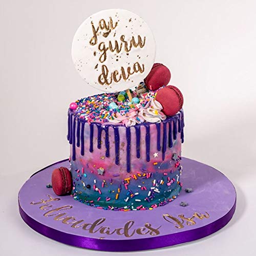 The image shows a colorful, multi-layered cake with a vibrant design featuring purple, pink, and blue icing. The cake has decorative elements like sprinkles, small colorful macarons, and edible decorations. At the top, there is a round cake topper with gold lettering that says "jai guru deva." The cake is displayed on a round purple cake board that has the words "Felicitades [name]" written in gold.