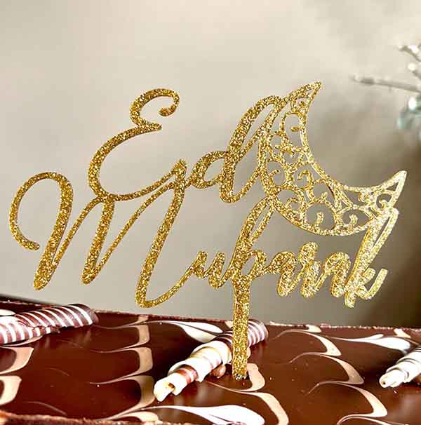 The image features a gold glitter cardstock cake topper that reads "Eid Mubarak." The lettering is stylized, with an ornate crescent moon design integrated into the text. The topper is displayed atop a chocolate cake, which is decorated with red and white striped peppermint candies. The background suggests a festive setting, enhancing the celebratory theme of Eid.