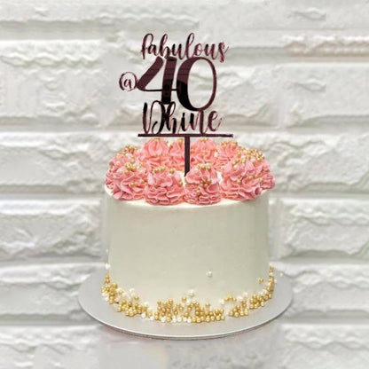 A white birthday cake with pink frosting swirls on top and decorated with small golden beads. It features a customized cake topper that reads "fabulous @ 40" in elegant script. The background has a textured white wall.