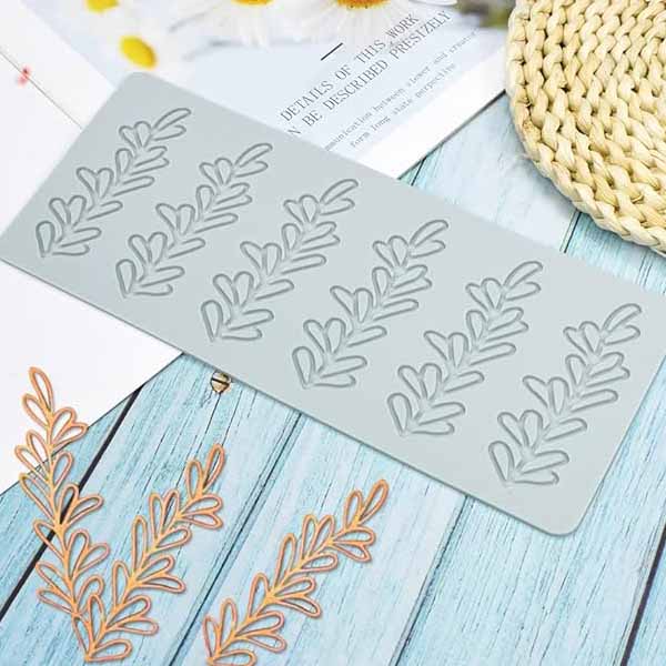 An image of a fine leaves silicone lace mould set against a light wooden background. The mould is rectangular, made of flexible silicone, and features intricate leaf patterns. Surrounding the mould are several small, orange, leaf-shaped embellishments, and parts of a woven circular mat and some white paper with printed text are partially visible in the background.
