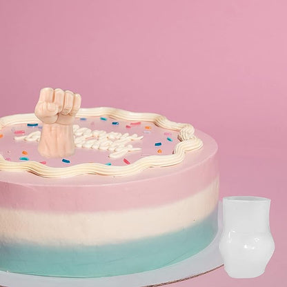 A colorful three-layered cake with pink, white, and teal frosting sits against a pink background. Atop the cake is a small, pink decorative fist made from silicone, surrounded by white decorations and colorful sprinkles. Next to the cake is a small, white, opaque cup.