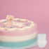 A colorful three-layered cake with pink, white, and teal frosting sits against a pink background. Atop the cake is a small, pink decorative fist made from silicone, surrounded by white decorations and colorful sprinkles. Next to the cake is a small, white, opaque cup.