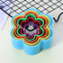 A flower cookie cutter set consisting of six colorful plastic cutters, stacked inside each other. The outermost cutter is light blue, followed by yellow, red, green, purple, and a small black cutter at the center. The background features a wire cooling rack and a light-colored surface.