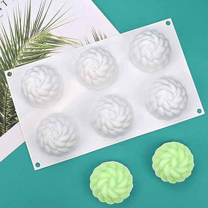A white silicone mold shaped like chrysanthemum flowers, featuring six cavities, rests on a teal surface. Two finished flower-shaped items, one green and one light green, are placed nearby. A tropical leaf design is visible in the background.