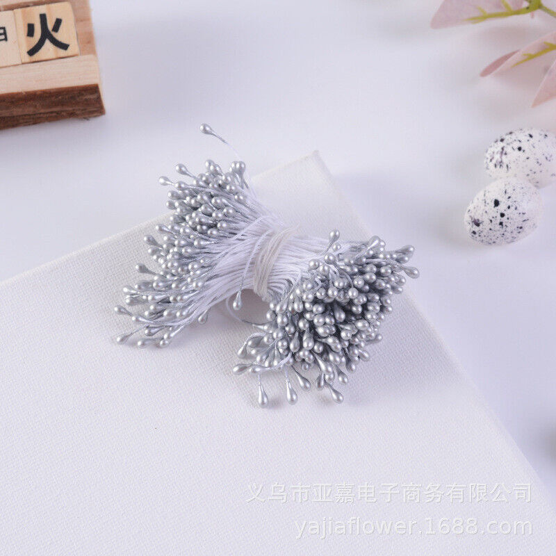 A bundle of flower stamens featuring approximately 70 silver-colored stamens attached to thin white thread. The bundle is laid on a white surface, with some decorative speckled eggs and a floral element slightly visible in the background.