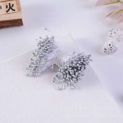 A bundle of flower stamens featuring approximately 70 silver-colored stamens attached to thin white thread. The bundle is laid on a white surface, with some decorative speckled eggs and a floral element slightly visible in the background.