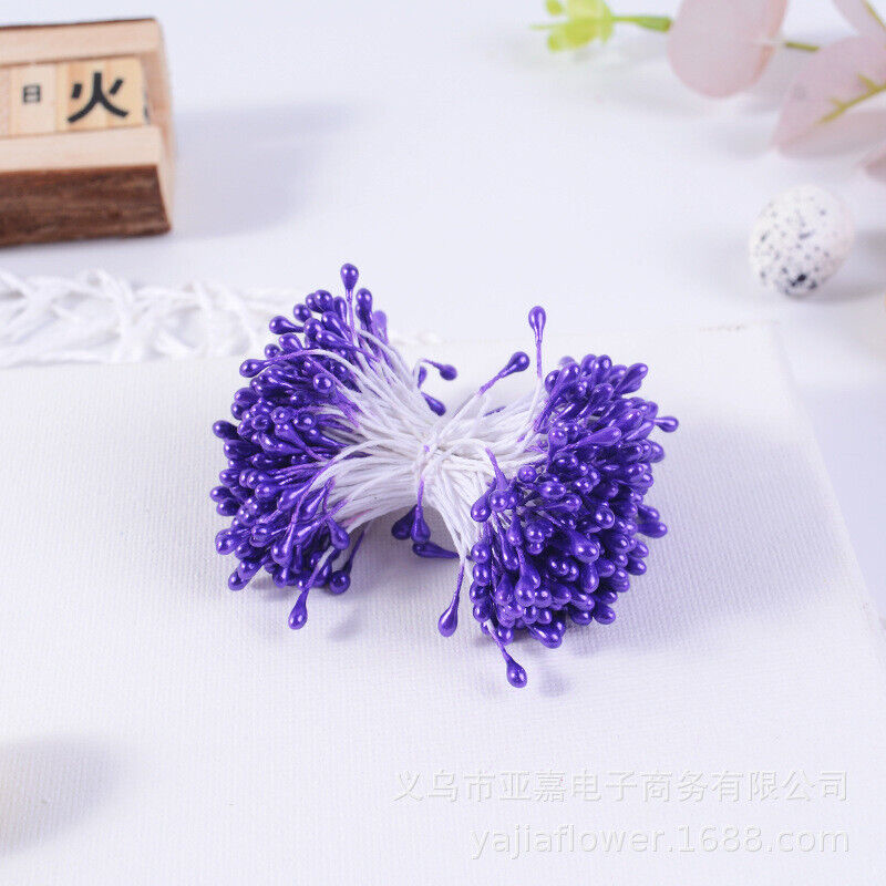 A bundle of decorative flower stamens is shown, featuring 70 individual stamens with purple teardrop-shaped tips. The stamens are clustered together, secured with white threads, and are placed against a light-colored background. Other floral decor elements can be faintly seen in the background.