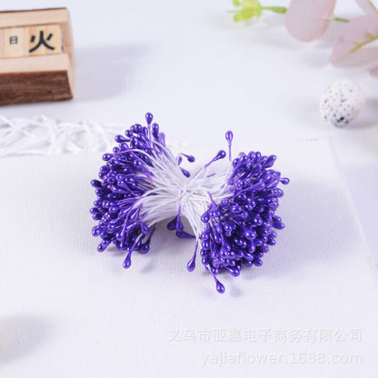 A bundle of decorative flower stamens is shown, featuring 70 individual stamens with purple teardrop-shaped tips. The stamens are clustered together, secured with white threads, and are placed against a light-colored background. Other floral decor elements can be faintly seen in the background.