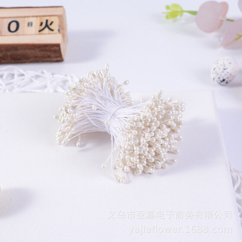 A bundle of flower stamens is displayed, featuring approximately 70 white stamens with small pearl-like tips, tied together with a thin white string. The background is a light-colored surface, and additional decorative elements, such as a wooden block and soft foliage, are faintly visible. The overall composition is clean and minimalistic, emphasizing the floral crafting product.