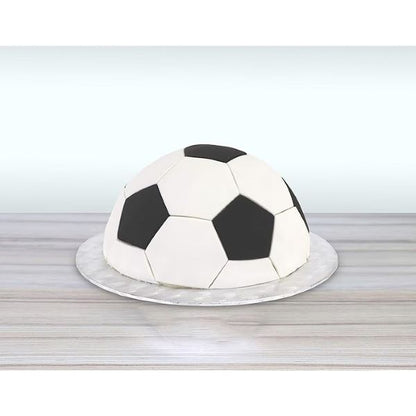 A round cake covered in fondant, designed to resemble a soccer ball. The cake features a white base with black pentagonal patches, mimicking the traditional pattern of a soccer ball. It is placed on a circular, shiny silver cake board, set against a light-colored background. The overall presentation highlights the cake&