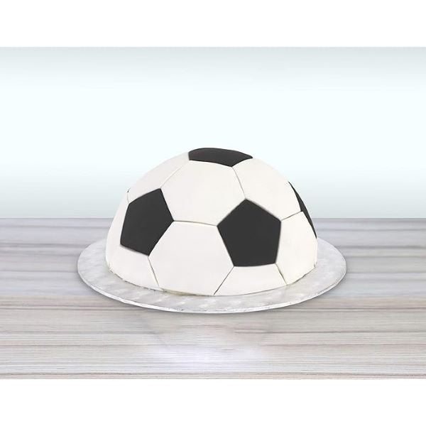 A round cake covered in fondant, designed to resemble a soccer ball. The cake features a white base with black pentagonal patches, mimicking the traditional pattern of a soccer ball. It is placed on a circular, shiny silver cake board, set against a light-colored background. The overall presentation highlights the cake&