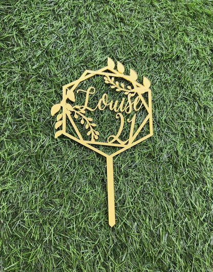 A golden acrylic cake topper displayed on a green artificial grass background. The topper features a geometric frame adorned with leaves and flowers, with the text "Louise" and the number "21" artistically integrated within the design. The topper&