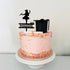 A round cake with pink frosting, decorated with colorful sprinkles around the bottom edge. Atop the cake is a black acrylic cake topper featuring a silhouette of a girl wearing a graduation cap and holding a diploma, along with the name "Maggie" and the year "2023." The cake sits on a white decorative cake stand.