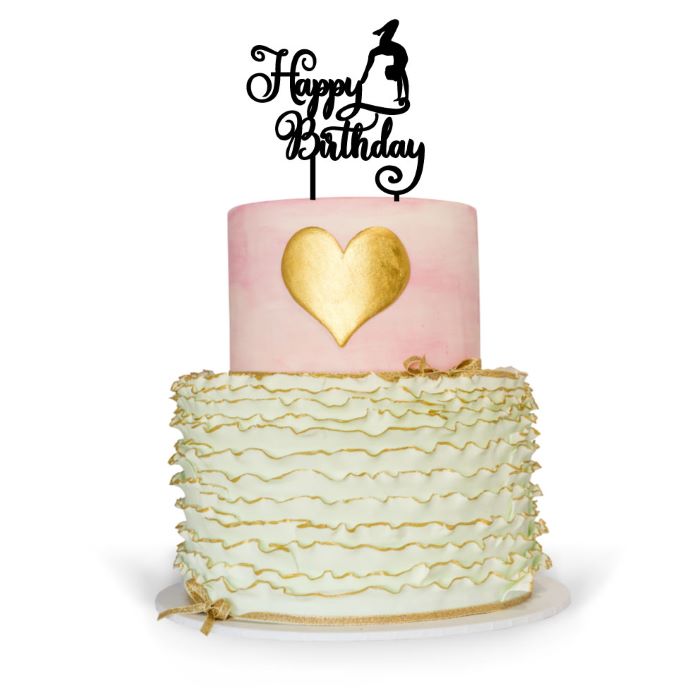 A two-tiered birthday cake featuring a pink top layer with a gold heart design and a white bottom layer decorated with ruffled cream icing. Topping the cake is a black cake topper that reads &quot;Happy Birthday&quot; with a gymnastics figure. The cake is placed on a round white base.