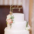 A two-tier white cake topped with a personalized acrylic cake topper that reads "Happy 21st Katelyn." The cake is decorated with delicate pink and white flowers, along with green leaves, adding a touch of elegance to the design. The background is softly blurred, highlighting the cake.