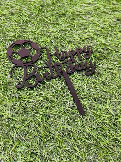A cake topper featuring the phrase "Happy Birthday" in an elegant script. It is designed with a soccer ball at the beginning of the text. The topper is placed on a green grass-like background, emphasizing its decorative purpose for a birthday celebration.