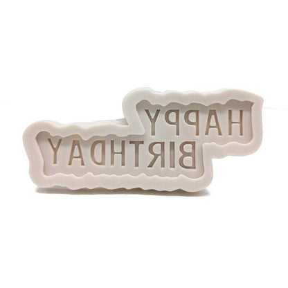 The image shows a silicone mold designed to create cake decorations. The mold features the phrase "HAPPY BIRTHDAY" in a bold, stylized typeface. The letters are indented and have a scalloped edge, giving them a decorative appearance. The mold is pale in color, likely light beige or white, and has a flexible, soft texture typical of silicone materials. The background is plain and white, emphasizing the mold&