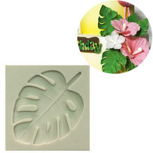 The image features a silicone mold shaped like a large tropical leaf, specifically a monstera leaf. The mold is a light color and displays detailed veining and a prominent central stem. In the corner of the image, there is a circular inset showcasing decorated flowers and leaves made from fondant or a similar material, used for cake decoration, suggesting a festive or tropical theme.