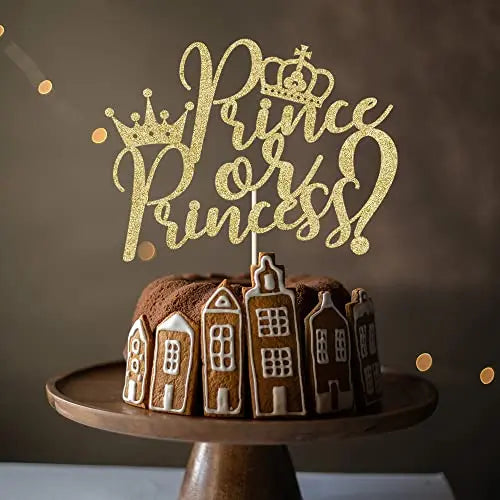 A decorative cake topper featuring the text &quot;Prince or Princess?&quot; in shiny gold script, topped with a crown. The topper is placed on a round chocolate cake decorated with gingerbread house shapes around the sides. The cake is set on a wooden cake stand, against a softly lit background.