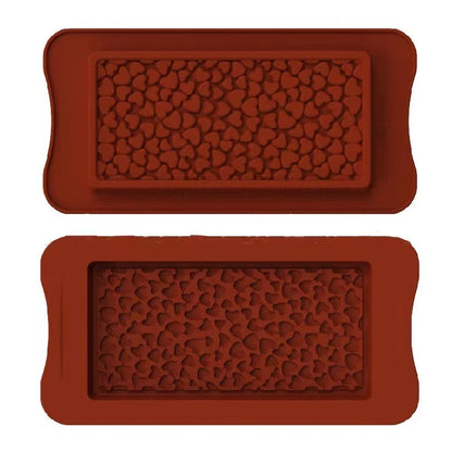 The image shows two silicone moulds for making chocolate bars, both designed with a pattern of small hearts. The moulds are rectangular in shape, with rounded edges and a textured heart design covering the surface. The top mould is positioned above the bottom mould, and both have a similar heart pattern, creating a decorative effect for the chocolate bars. The colour of the moulds is a rich, reddish-brown.