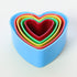 The image shows a set of five heart-shaped cookie cutters stacked inside one another. The outermost cutter is a light blue color, followed by a red cutter, a green cutter, a yellow cutter, and finally a smaller pink cutter in the center. The cutters are made of a flexible material and have a smooth finish. They are arranged in a way that highlights their different sizes while maintaining a heart shape. The background is simple and light, emphasizing the colors of the cookie cutters.