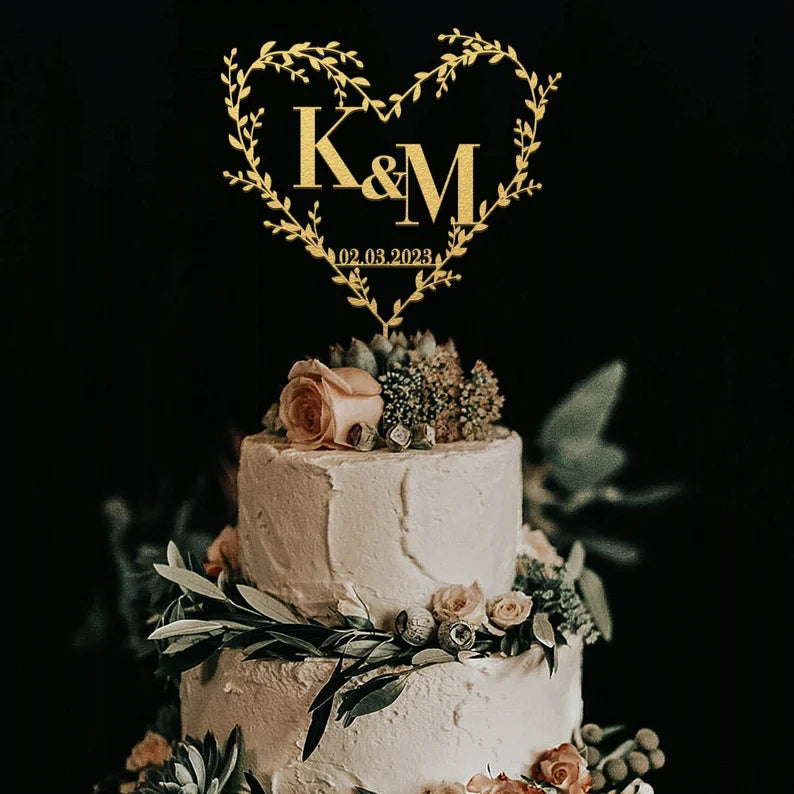 The image features a three-tiered wedding cake adorned with a heart-shaped acrylic cake topper. The topper is designed with the initials "K &amp; M" and the date "02.03.2023," surrounded by delicate greenery. The cake itself has a textured white frosting and is decorated with light pink roses and various greenery elements on the top and around the base. The background is dark, providing contrast to the cake and the topper.