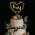 The image features a three-tiered wedding cake adorned with a heart-shaped acrylic cake topper. The topper is designed with the initials "K & M" and the date "02.03.2023," surrounded by delicate greenery. The cake itself has a textured white frosting and is decorated with light pink roses and various greenery elements on the top and around the base. The background is dark, providing contrast to the cake and the topper.