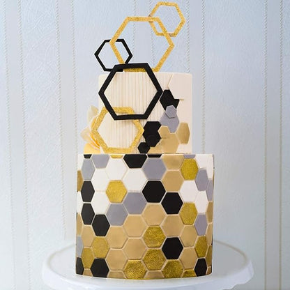 A decorative cake featuring hexagon patterns in shades of black, gold, and gray. The cake is adorned with hexagon-shaped toppers in gold glitter and a black hexagon design. The background is a subtle striped pattern, and the cake is placed on a white pedestal.