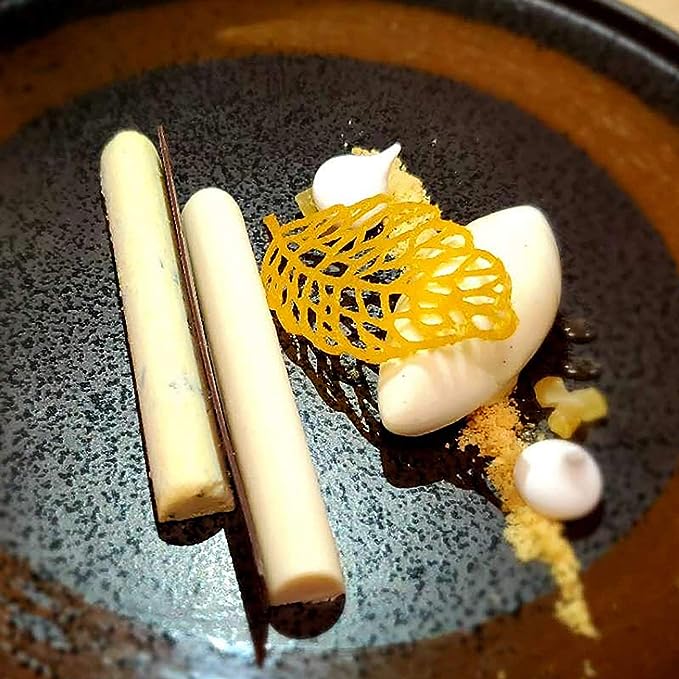 A black plate featuring an artistic dessert presentation. On the plate are two white cylindrical shapes, a dark chocolate-like stick, a white scoop of cream, and a delicate yellow crunchy structure resembling a leaf. Small dots of a white sauce and some golden crumb are scattered around the dessert.