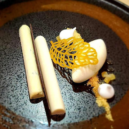 A black plate featuring an artistic dessert presentation. On the plate are two white cylindrical shapes, a dark chocolate-like stick, a white scoop of cream, and a delicate yellow crunchy structure resembling a leaf. Small dots of a white sauce and some golden crumb are scattered around the dessert.