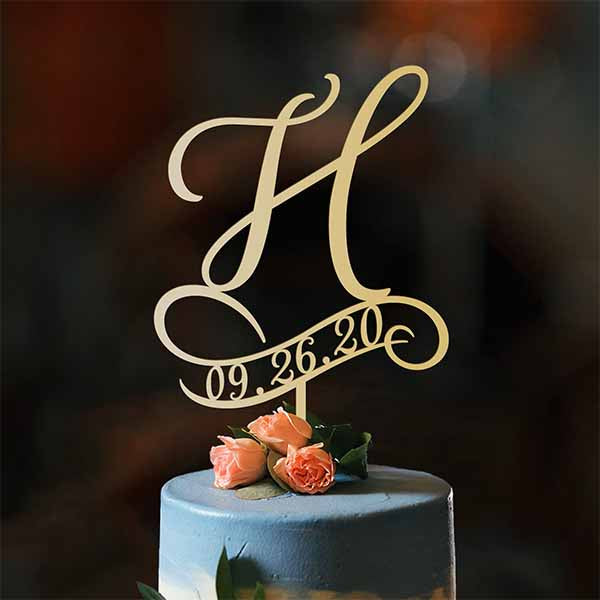 An elegant acrylic cake topper featuring a large, stylized initial &quot;H&quot; in gold, accompanied by a decorative swoosh design below it. The date &quot;09.26.20&quot; is elegantly incorporated into the design beneath the initial. The topper is placed on a tiered cake adorned with soft blue icing and topped with three peach-colored roses, adding a touch of floral decoration. The background is softly blurred, emphasizing the cake and topper.