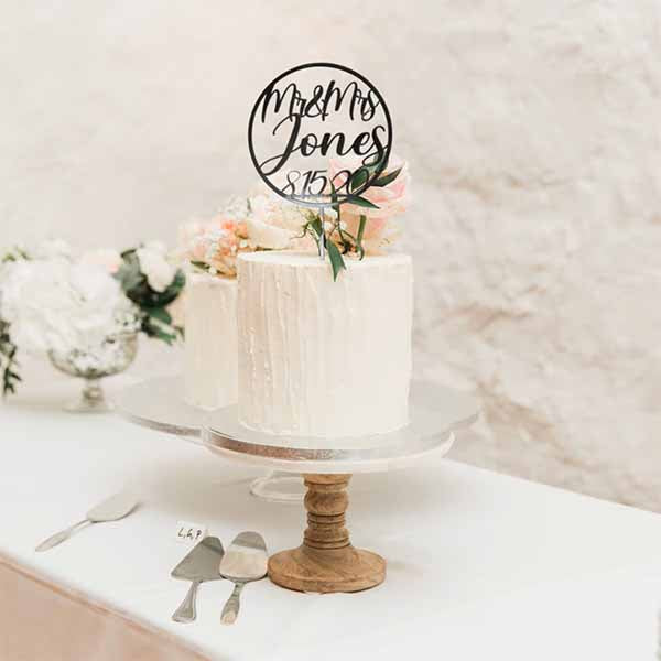 A wedding cake displayed on a round silver cake stand features a modern acrylic cake topper that reads &quot;Mr &amp; Mrs Jones&quot; with the date &quot;81520&quot; inside a circular design. The cake is decorated with a smooth white icing, and there are small flowers and greenery on top. In the background, a second cake is partially visible, along with two serving utensils placed beside the cakes on a light-colored tablecloth.