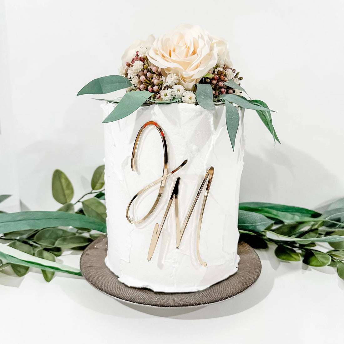 A decorated white cake topped with artificial flowers, including a light-colored rose and greenery. The cake features a shiny gold acrylic charm with the initials "JM" on its side. The cake sits on a round brown cake stand, surrounded by greenery. The background is a simple light color.