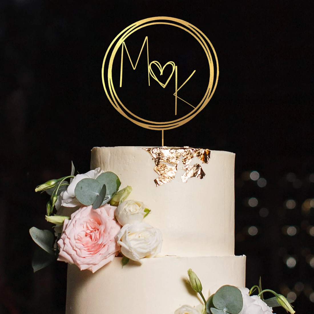 A three-tiered white wedding cake topped with a gold acrylic cake topper featuring the initials "M" and "K" inside a circular design with a heart in between. The cake is adorned with delicate pink and white flowers, as well as eucalyptus leaves, and has gold leaf accents on the top tier. The background is dark, highlighting the cake and its decorations.
