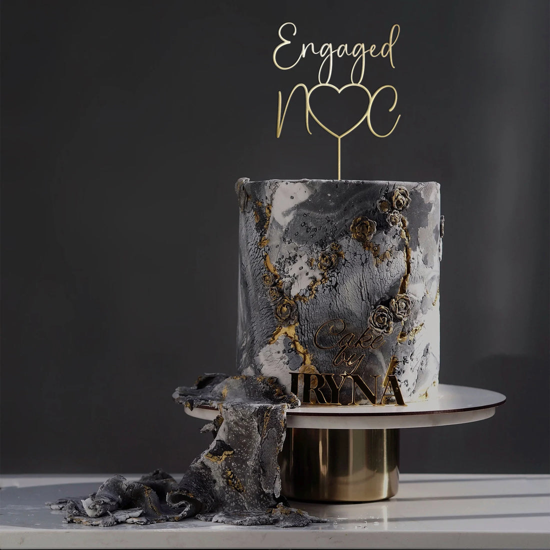 A two-tiered cake featuring a marbled gray and white design with gold accents and floral decorations. Topping the cake is a personalized acrylic cake topper that reads "Engaged" with a heart, displayed on a circular cake stand with a gold base. A piece of cake has been cut, showing the inside. The background is dark, enhancing the elegant presentation of the cake.